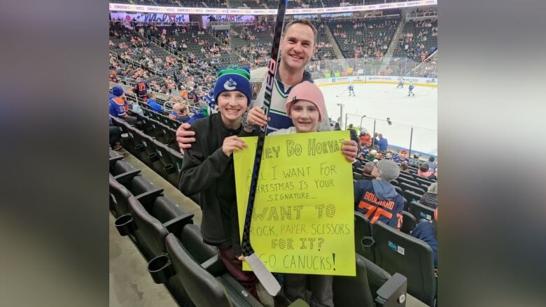 On the spectators' platform inside an arena, a man poses with two boys who are holding a hockey stick and a big yellow piece of paper with words.