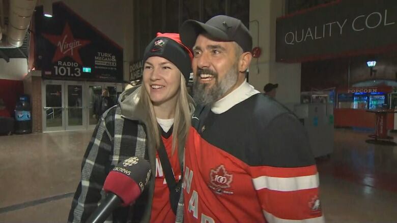 A man and a woman wearing Team Canada jersey smile while talking to reporter. 