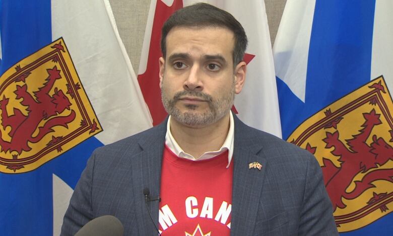 A man with a gray beard and short dark hair stands in front of the Nova Scotia and Canada flags.