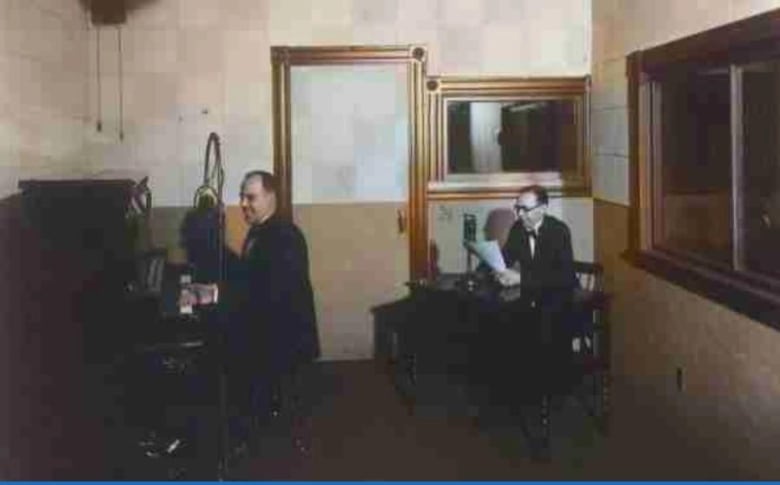 A phot of a small room with two windows. A man is seated at a table, a microphone in front of him, holding papers in his hand. A second man is seated at a piano.