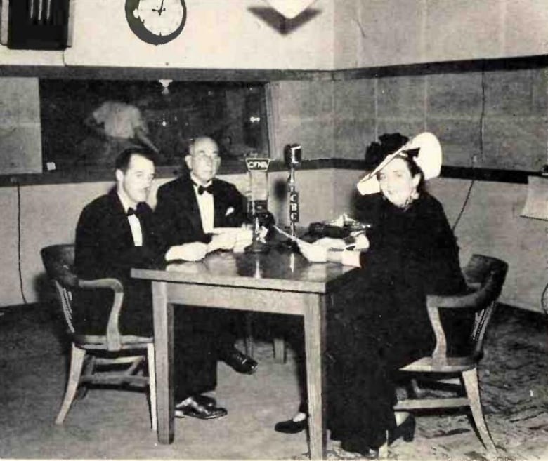 A woman and two men sit around a table in a radio studio. The men wear suits, the woman is in a dress and a large hat. 