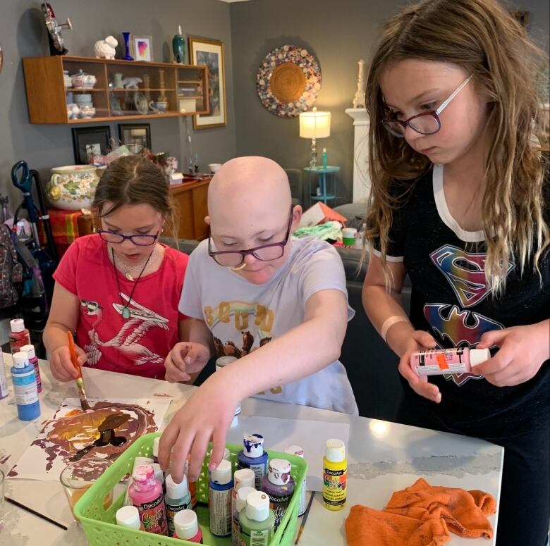 Three girls sit around a table looking at different paints. One paints a picture on a piece of paper.