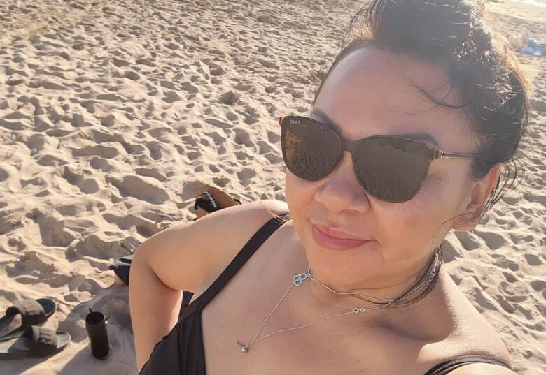 A woman wearing sunglasses stands on a sandy beach and smiles towards the camera.