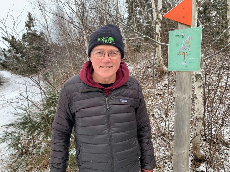 A man wearing a puffed jacket and an Island Trails hat stands near a trail marker with an orange flag, smiling at the camera.