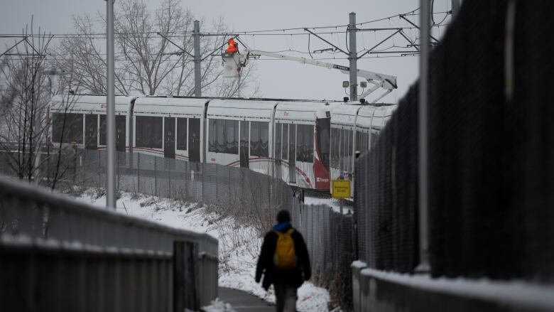 A worker in an orange jacket works to repair wires above a stalled light rail train as a person walks by in the foreground.