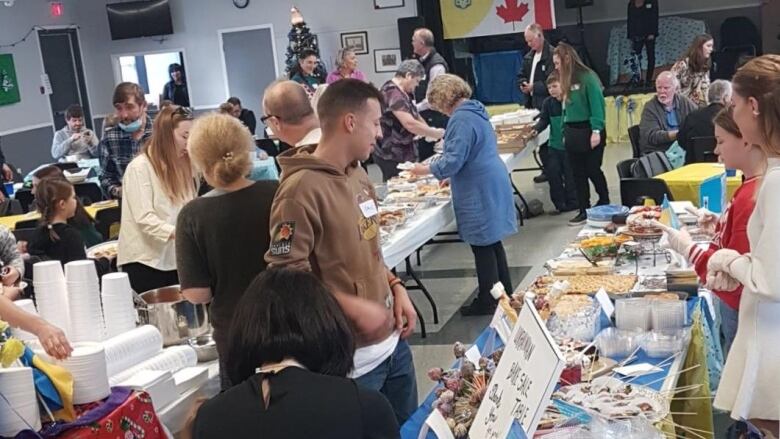 A number of people stand near long tables filled with food and gift items. Ukrainian and Canadian flags are in the background.