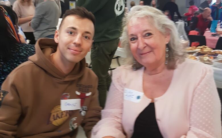 Young man in brown hoodie on the left sits next to woman in pink jacket on the right.