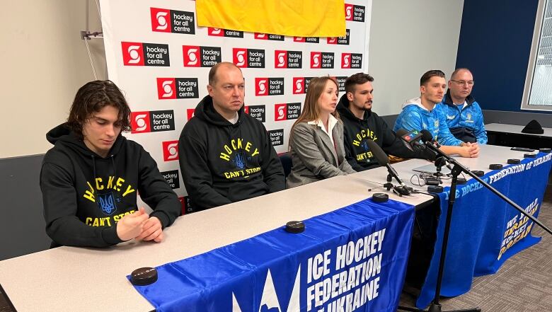 Six people sit on one side of a table. A woman in the middle speaks into a microphone.