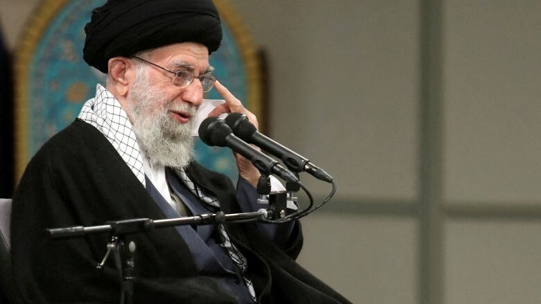 A man in black robes and a black turban sits on a stage in front of microphones.