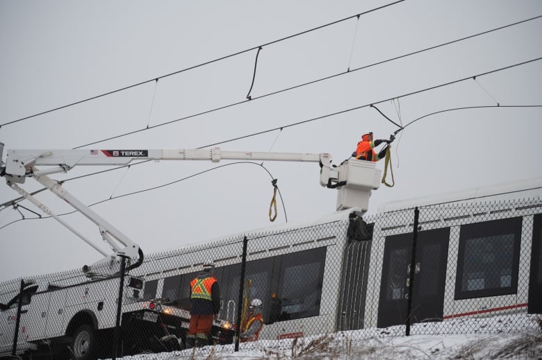 A worker in a bucket truck works on power cables above a light rail train.