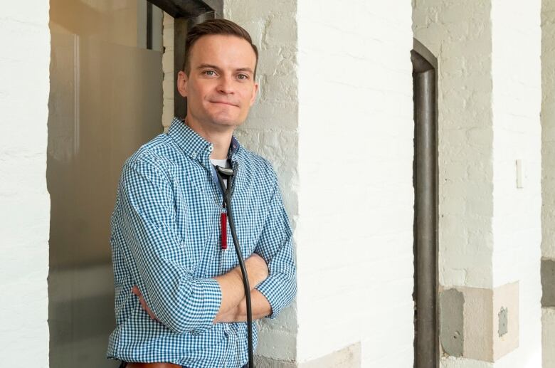 A man in a blue and white checkered dress shirt smiles with his arms folded.