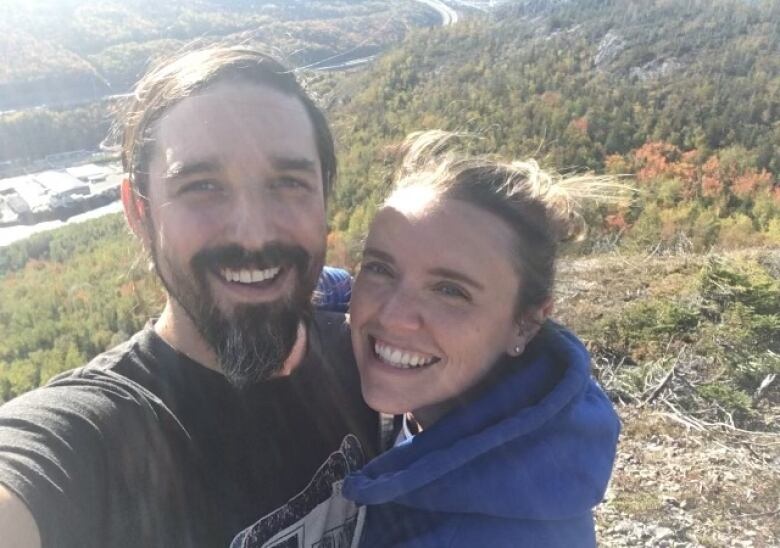 A man and woman smile together on top of a hill covered in trees.