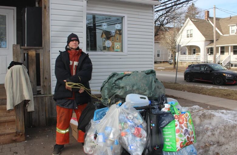 man dressed in layers standing with a shopping cart overflowing with garbage bags filled with cans and bottle and a tarp.