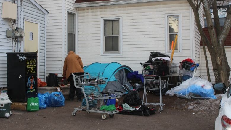 A man walking around a parking lot behind a building with a tent, several shopping carts and bags of belongings scattered around 