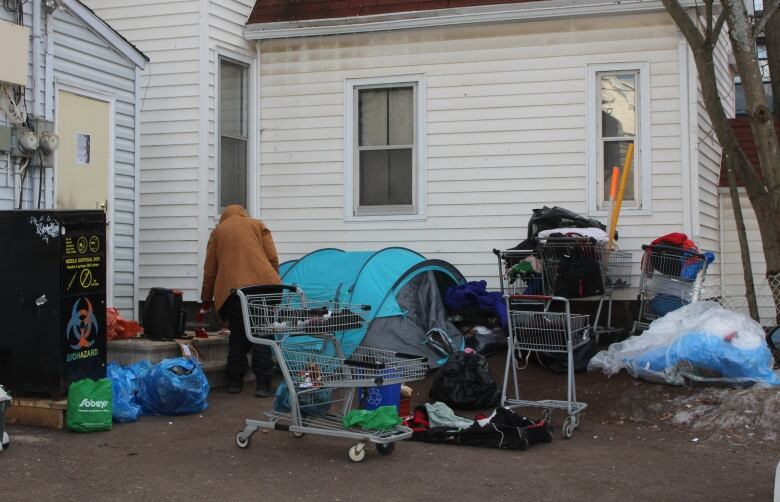 A man walking around a parking lot behind a building with a tent, several shopping carts and bags of belongings scattered around 