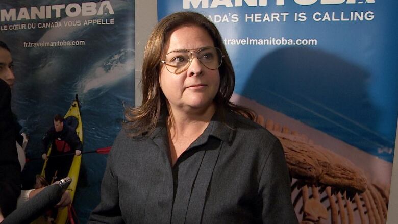 A woman with shoulder-length brown hair wearing aviators in a black button down shirt stands in front of a blue banner that says, 