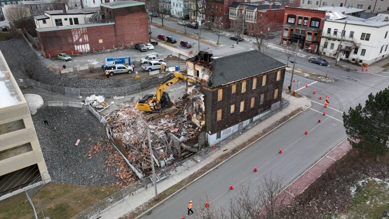 An aerial view of a brown wooden house being demolished by an excavator. A cross section of the house can be seen as the excavator tears away at it. 