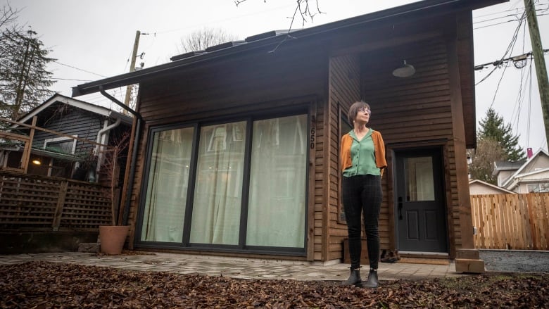 A woman stands outside a small house attached to a bigger one looking away from the camera.