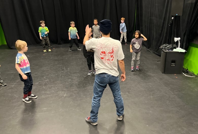 A man in a dance studio stands with his back to the camera while a group of young kids are spread out in front of him, watching. 