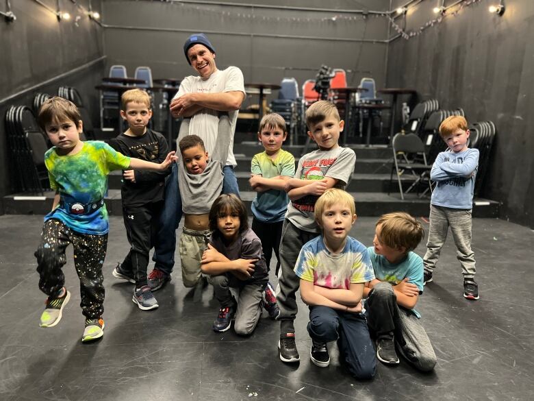 A group of young boys stand facing the camera in hip-hop dance poses inside a studio, with a smiling man standing and posing among them.