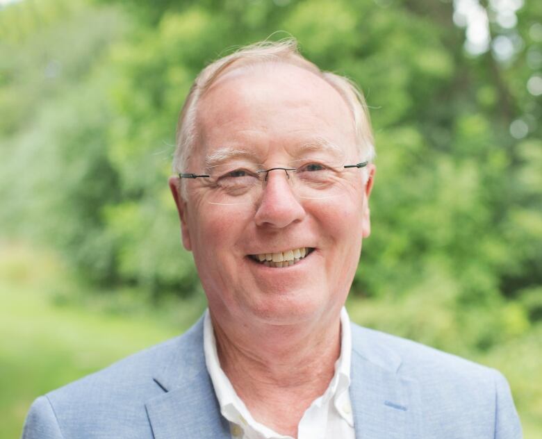 a man in a light blue suit jacket and glasses smiles outside in front of a tree