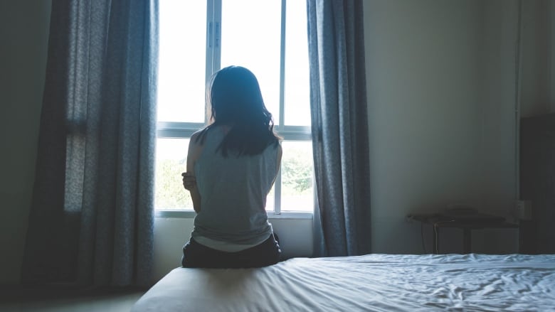 An unidentifiable woman sits in front of a window on a bed with her back to the camera.