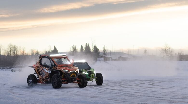 Two ATVs, one red and one green, slide on a frozen lake, kicking snow up into the air as the sun sets behind them. 