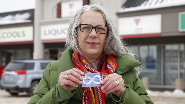 A woman with long grey hair wearing a green coat and a striped red scarf holds up a card that is blue with a white infinity symbol. She is standing in front of a liquor store.