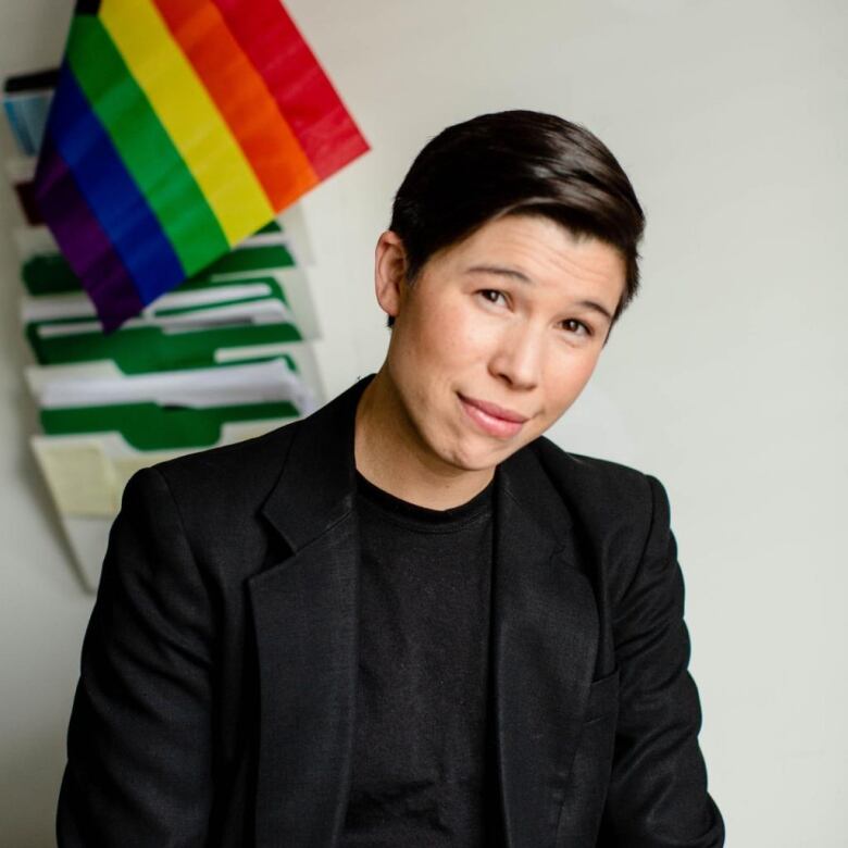 A person with short, dark hair and wearing a black shirt and jacket smiles at the camera.