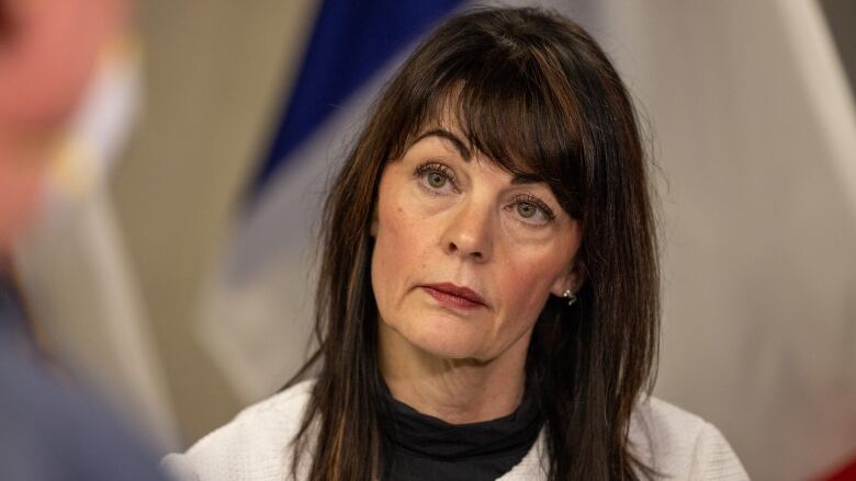 A woman with long hair is seated. She is wearing a white blazer with a black shirt underneath.