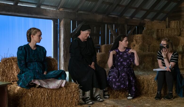 Four women sit on haystacks in a dark loft in a scene from the film Women Talking.