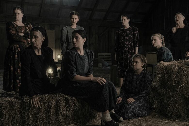 A group of eight women sit in a dimly lit hayloft with lanterns nearby