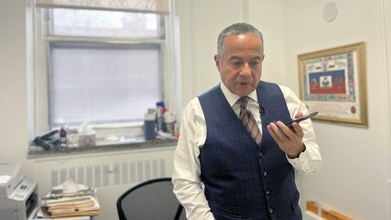 A man in a vest speaks into a phone inside a white office, with a Haitian flag on the wall.