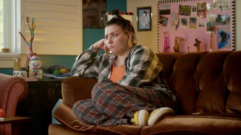 A woman sits on a couch in her pyjamas, with a concerned look on her face.