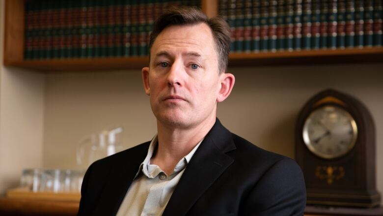 A slim man with short brown hair, wearing a collared shirt and black blazer, looks directly at the camera. There is a bookshelf behind him.