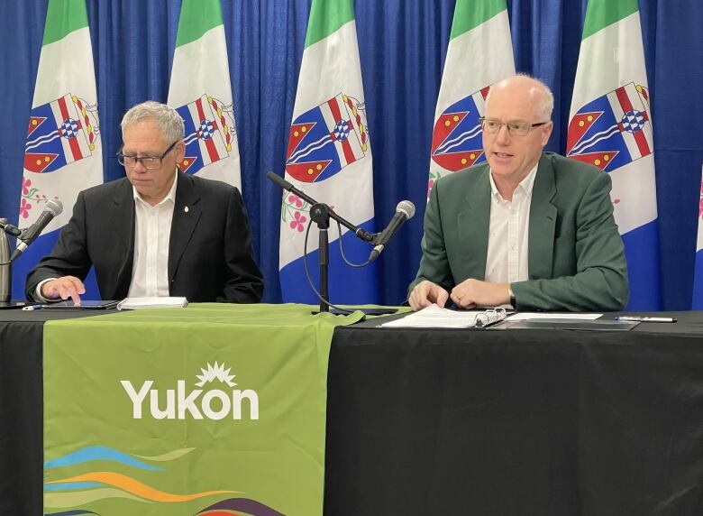 Two men sit at a table before microphones, with a 'Yukon' banner on the front of the table and a line of flags behind them.