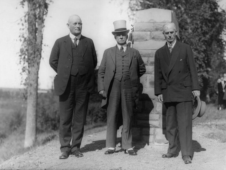 black and white photo of three men, one in a top hat, standing in front of stone monument