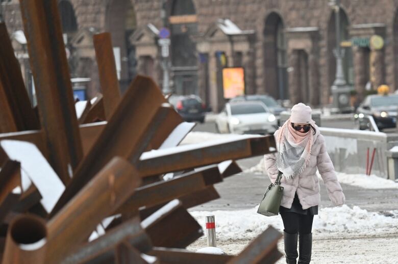 A woman wearing sunglasses, winter hat, scarf, coat and a purse walks along a snow-covered street. Buildings and cars are visible, and the streetscape would seem completely normal if not for the rusted pieces of metal meant to stop tanks.