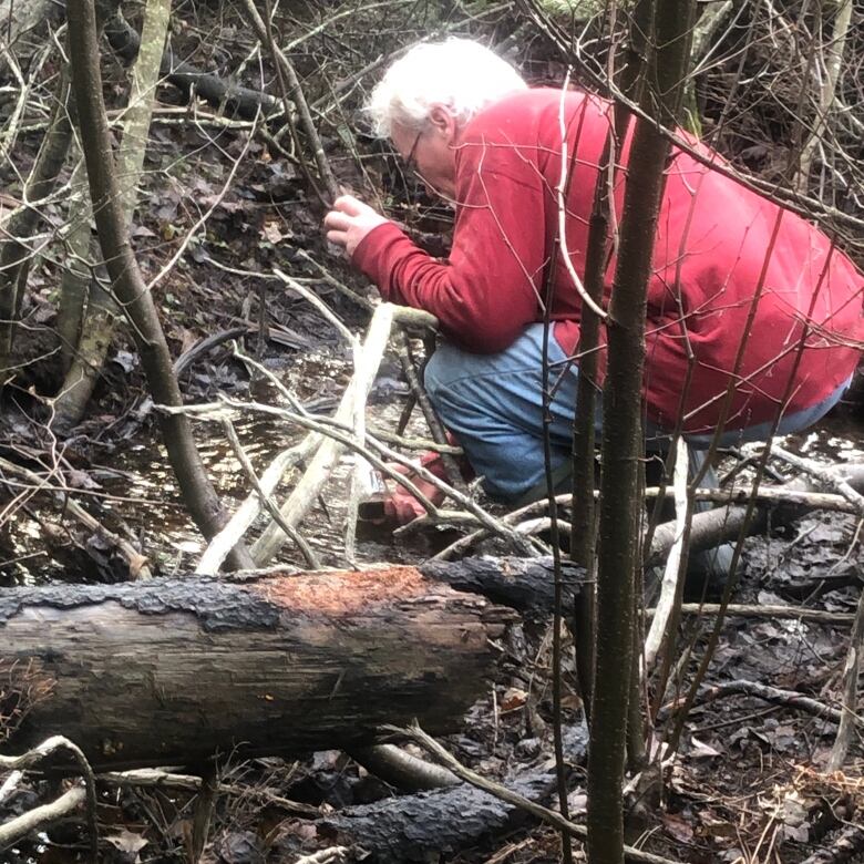 A man in a red coat is seen crouching by a stream in the forest.