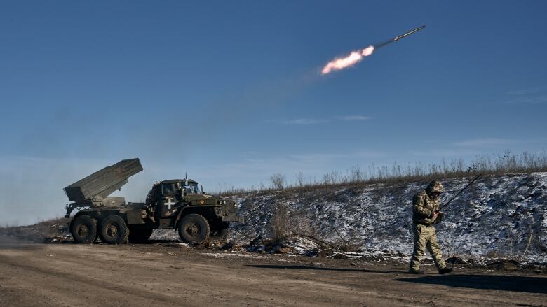 A truck with a rocket launcher on the back sits at the side of a road. A missile that has just been fired sails upward. A man in military uniform walks by the side of the road, head down.