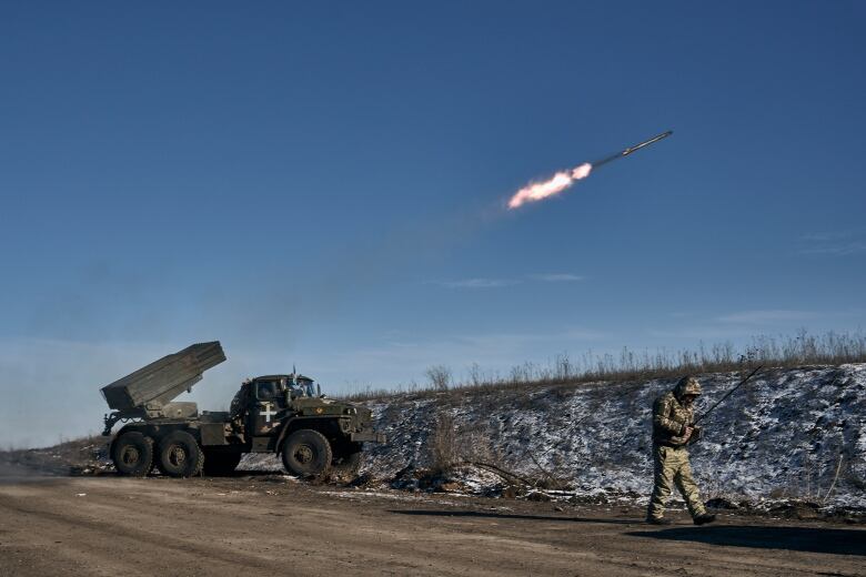 A truck with a rocket launcher on the back sits at the side of a road. A missile that has just been fired sails upward. A man in military uniform walks by the side of the road, head down.