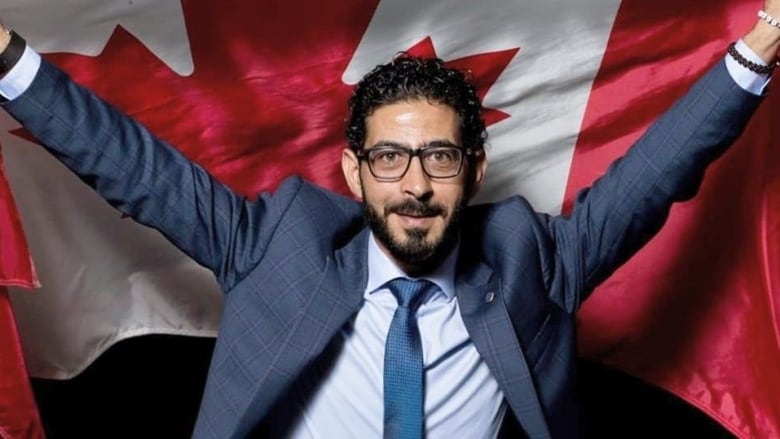 A man stands in a suit holding a Canadian flag behind him.