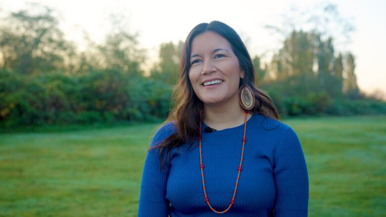 A smiling woman in a beaded necklace stands facing the camera with green trees behind her. 