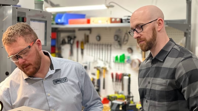 Two men wearing glasses inspect a machine used to make taps and faucets.  Both are bearded and the boss, who is slightly shorter, is wearing a button down company shirt with the name Tapmaster on it.  His worker is bald and wearing a  grey plaid shirt.  