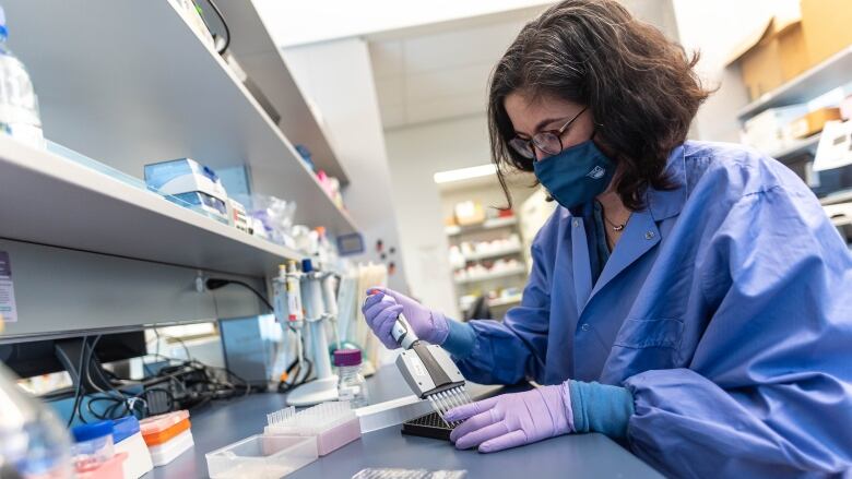 A woman in blue medical garb wearing a blue medical mask squeezes the contents of a vial into a device.