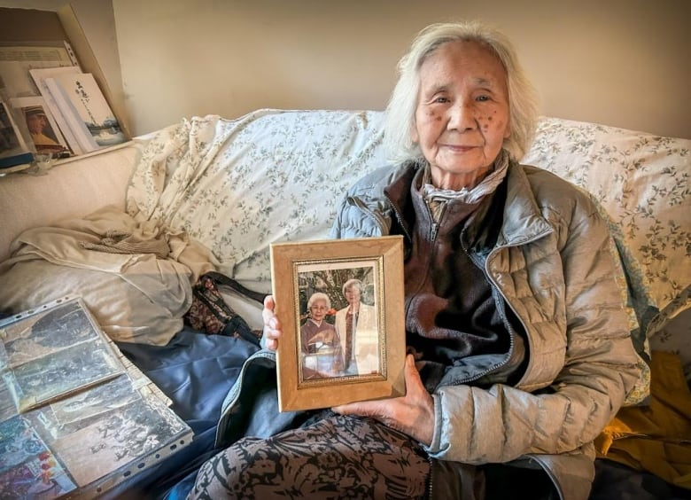 An old woman sits on a couch and shows a photo of herself from decades past with her mother.