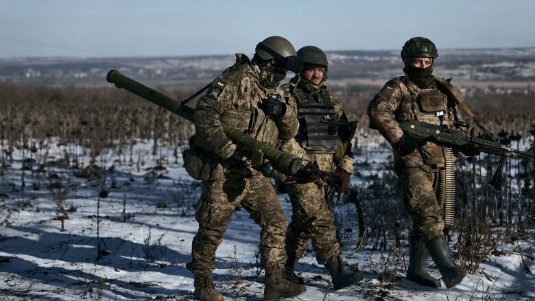 Three Ukrainian soldiers in army uniforms walk while carrying a rocket launcher and a machine gun.