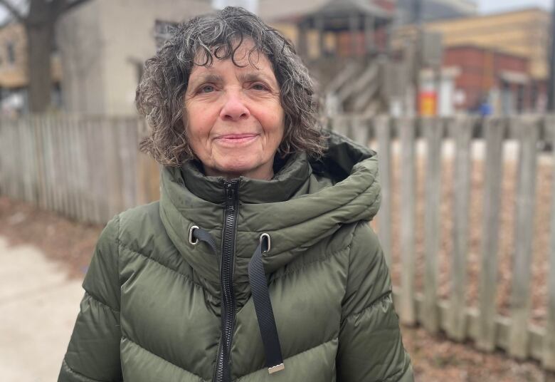 An older woman in an army green puffer coat stands outside next to a wooden fence. A playground structure and school buildings are seen behind her.