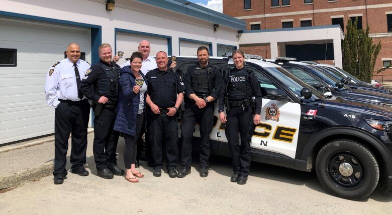 Several uniformed police officers stand in front of a police cruiser on a sunny day.