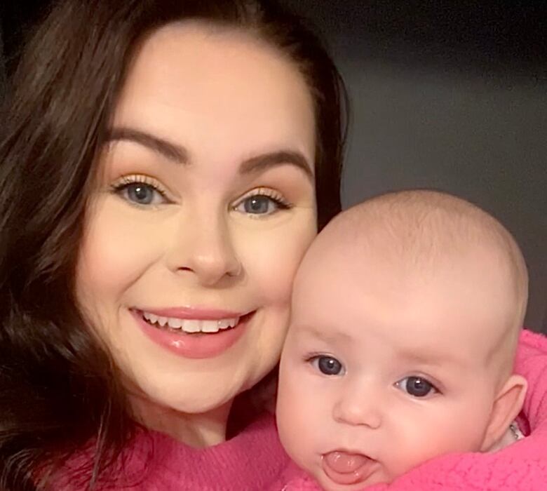 A smiling woman with long, brown hair holds her baby. The baby is wearing a pink sweater.